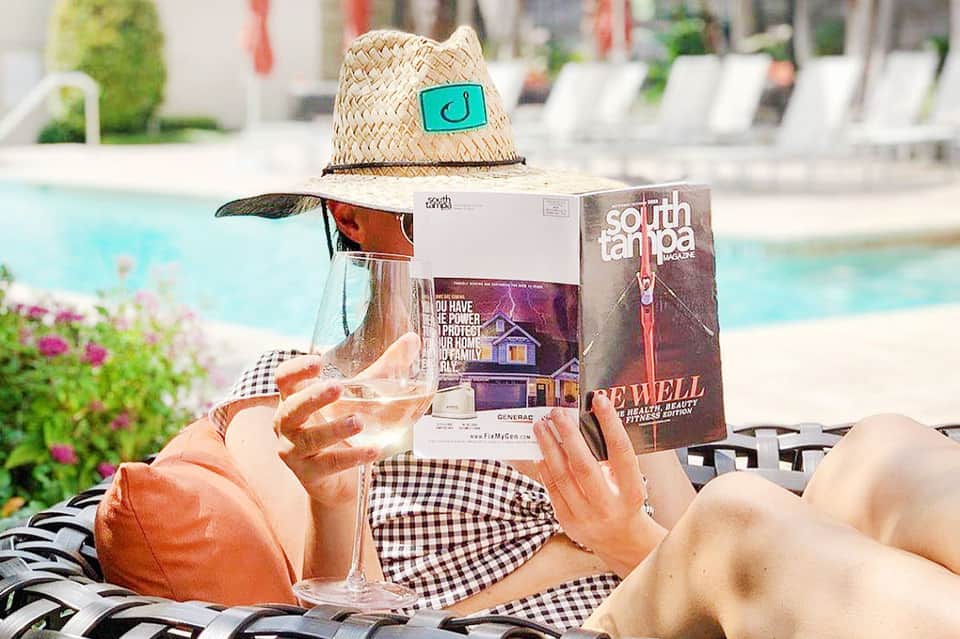 Woman in checkered outfit reading at poolside with straw hat and tropical drinks