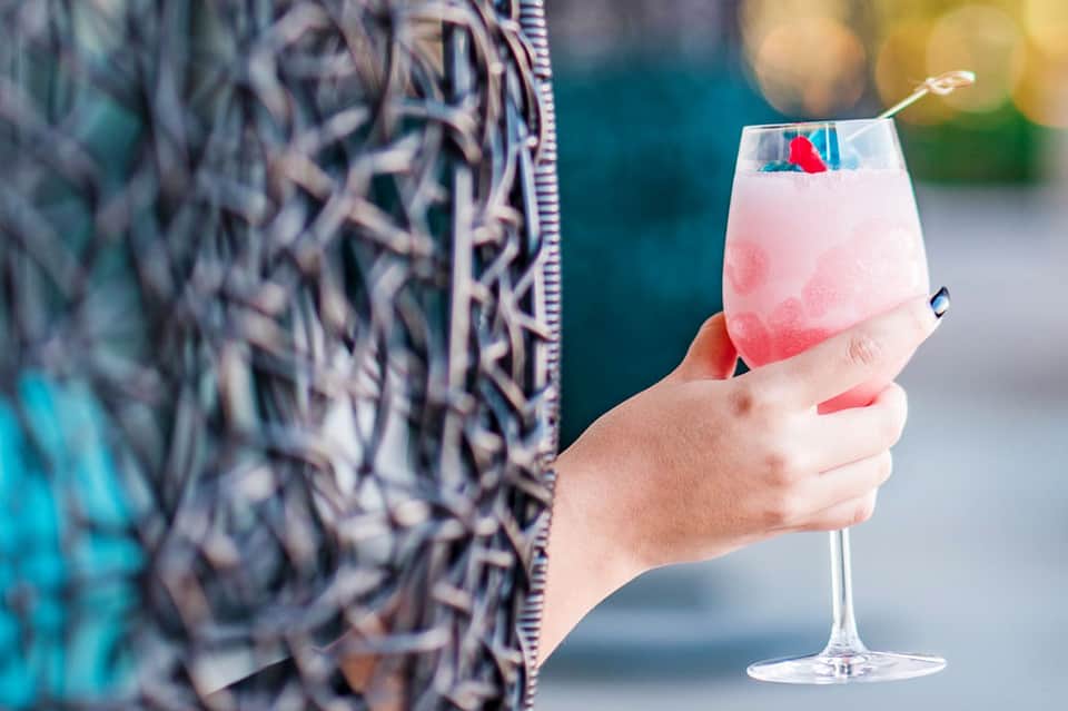 Hand holding pink cocktail with colorful flags, ocean and beach loungers blurred in background