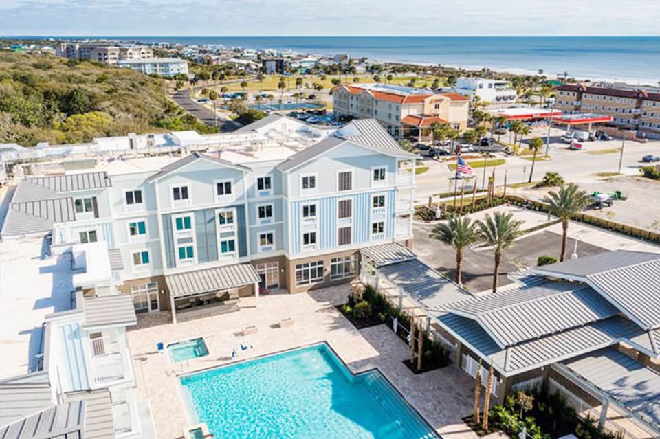 Aerial view of beachfront resort with pool, white buildings, palm trees, and ocean beyond