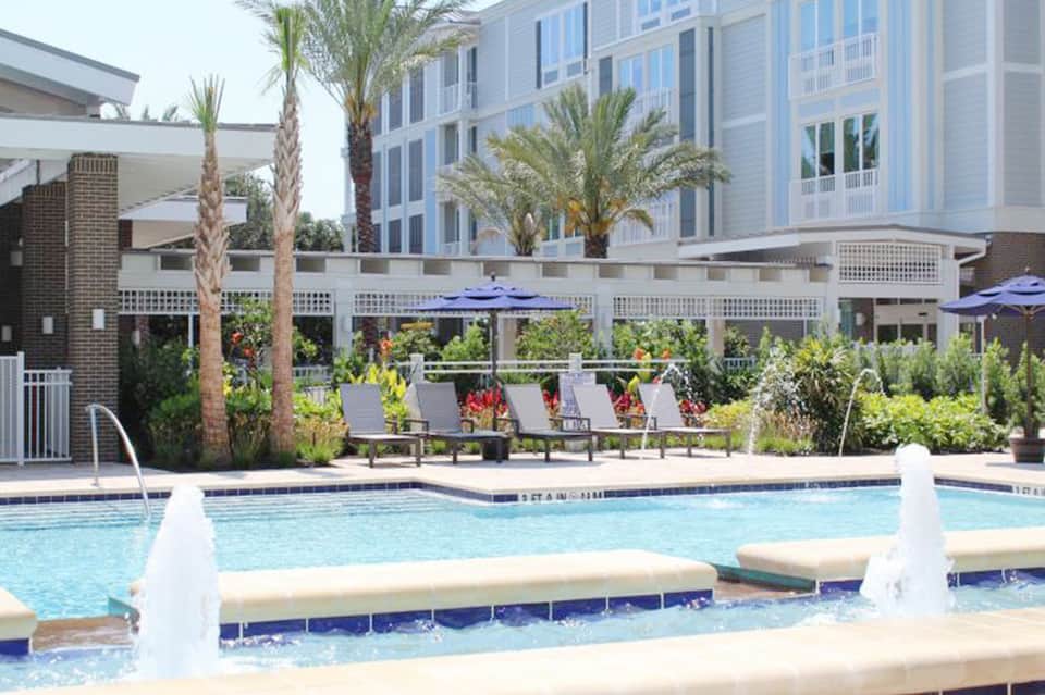 Resort pool area with lounge chairs, blue umbrellas, palm trees, and contemporary building backdrop