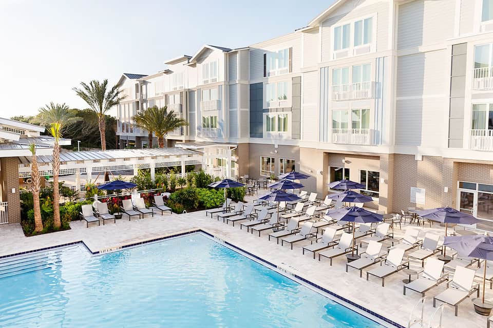 Modern resort pool area with multiple white umbrellas, lounge chairs, palm trees, and contemporary building