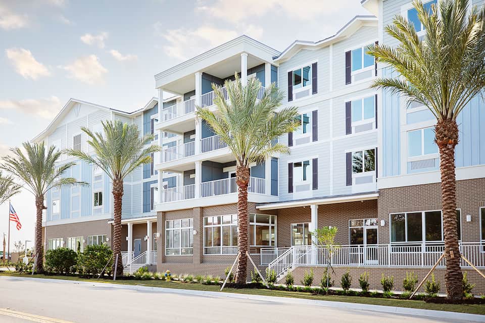 Coastal residential building with white and blue exterior, multiple balconies, palm trees, and ground-level retail space