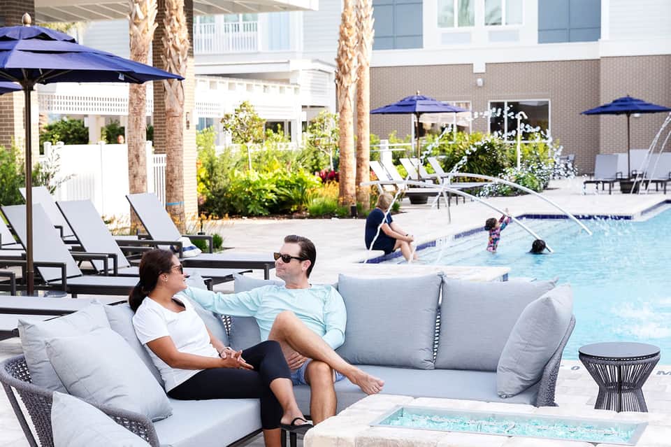 Couple relaxing on gray poolside seating with blue umbrellas and swimming guests in background
