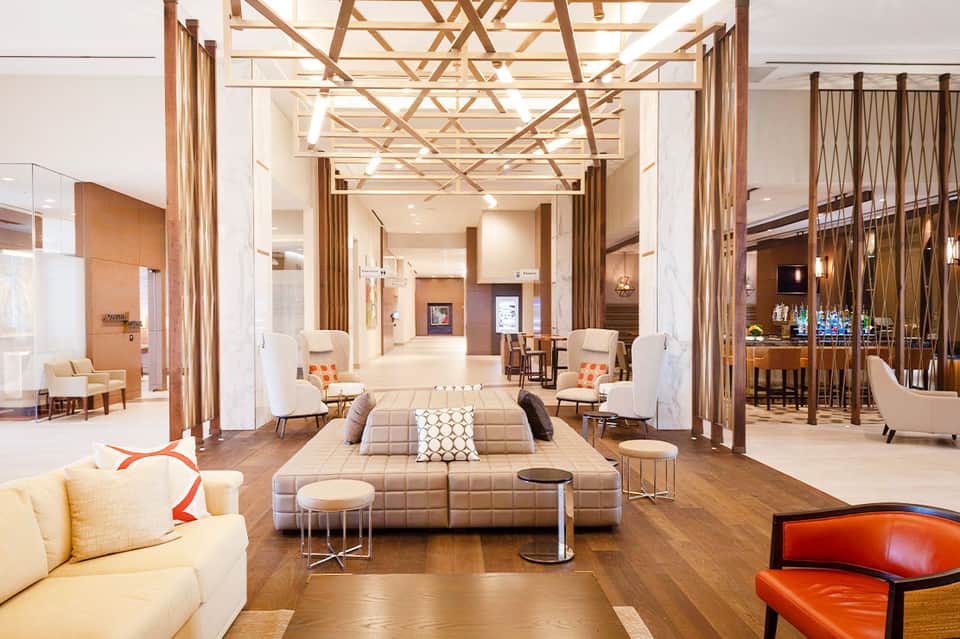 Modern hotel lobby with neutral tones, geometric ceiling, lounge seating, and bar area