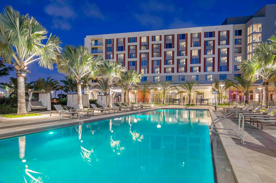 Illuminated resort pool at dusk with palm trees, lounge chairs, and modern brick hotel building
