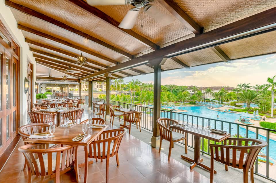 Elevated restaurant dining area with wooden beamed ceiling overlooking pool and palm trees