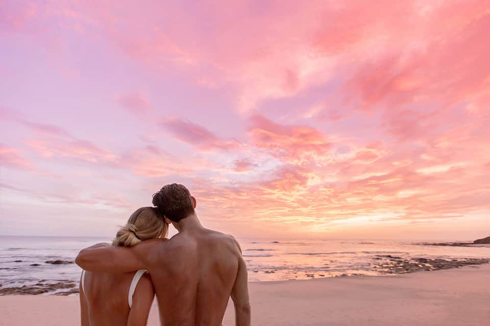 Couple embracing on beach at sunset with pink and orange sky over ocean waves