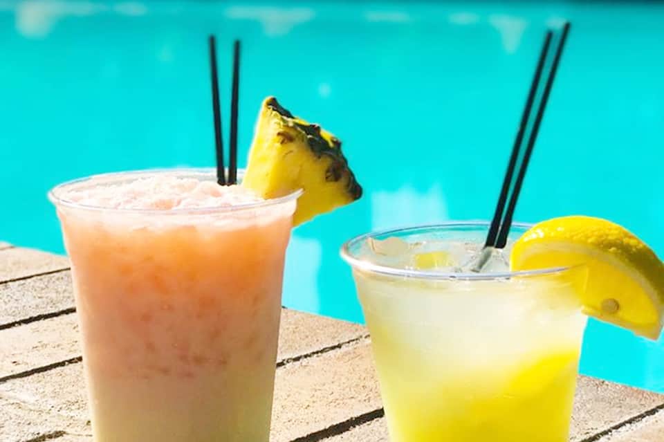 Two tropical cocktails by the pool: a peachy drink with pineapple garnish and a yellow citrus drink with lemon slice