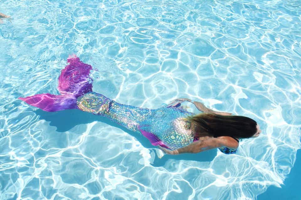 Woman swimming underwater in crystal-clear turquoise pool wearing colorful mermaid tail fin