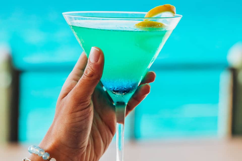 Hand holding a vibrant turquoise cocktail with lemon garnish against an ocean backdrop