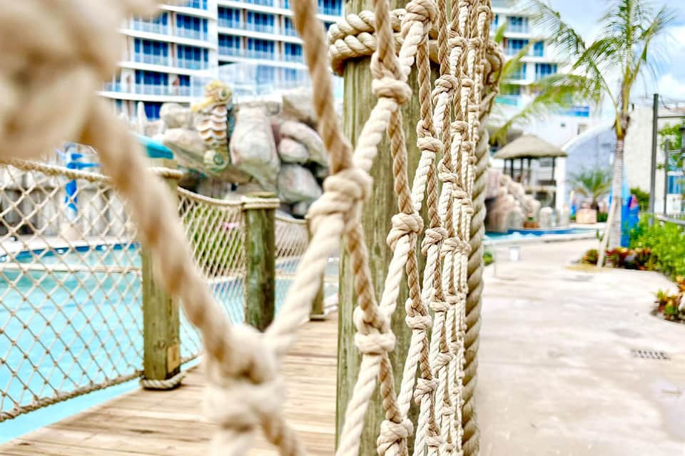 Rope railings on wooden deck leading to pool area with resort buildings in background