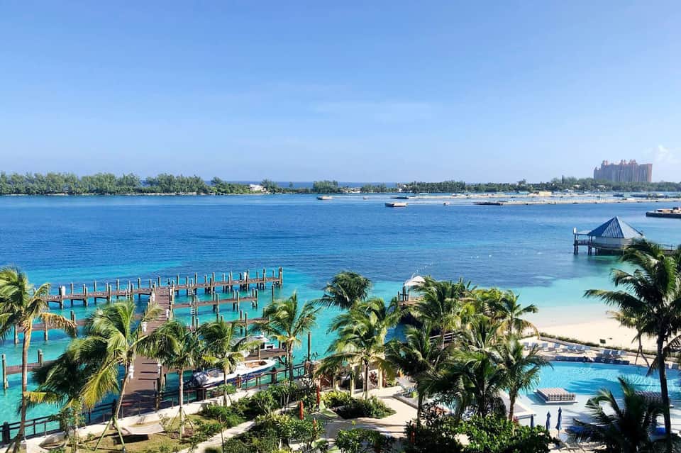 Tropical beach resort with wooden dock, palm trees, turquoise water, and distant hotel buildings