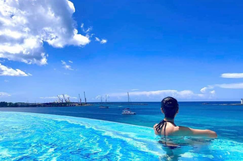 Woman swimming in infinity pool overlooking turquoise ocean waters with boats and industrial port