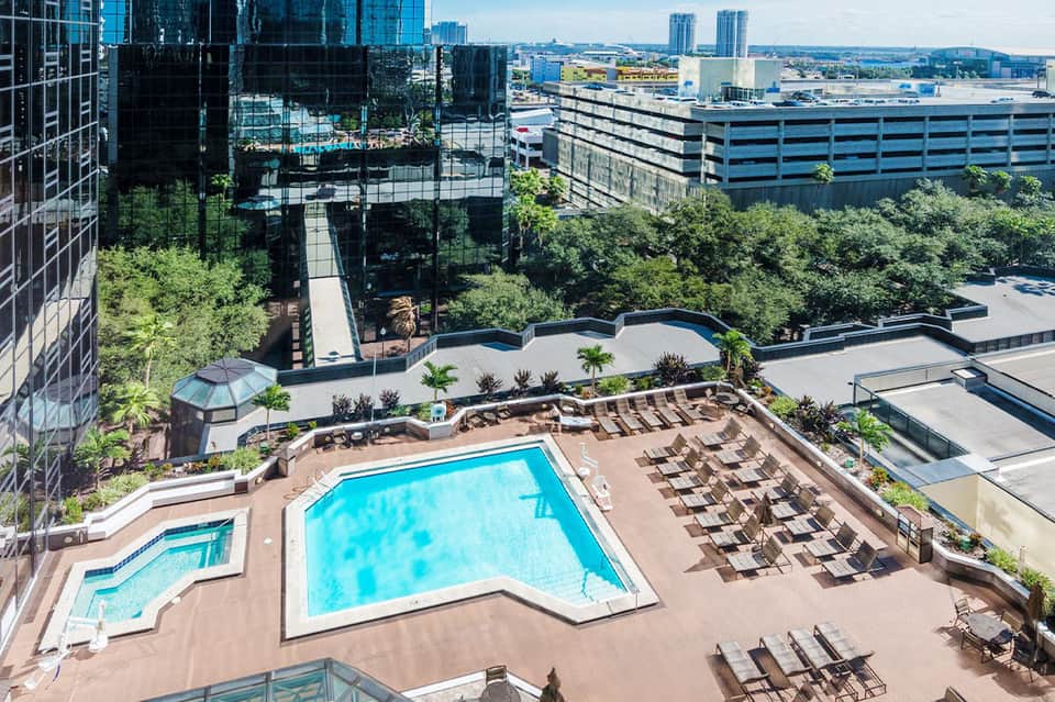 Aerial view of resort pool deck with lounge chairs, palm trees, and city skyline in background