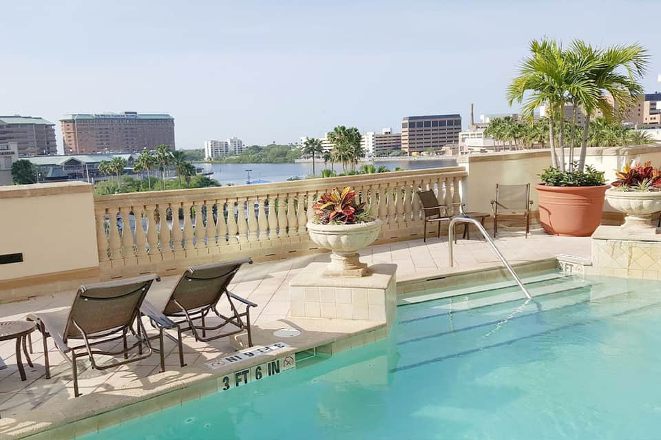 Waterfront pool terrace with lounge chairs, decorative planters, balustrade railing, and city skyline view