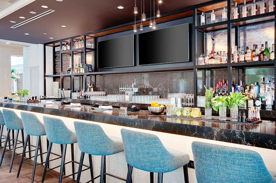 Modern hotel bar with turquoise upholstered seats, dark marble countertop, and illuminated shelving