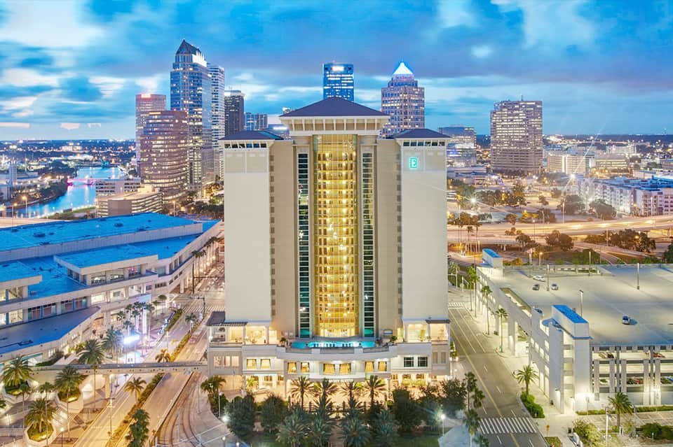 Tall hotel tower illuminated at dusk overlooking city skyline with blue pool facility visible