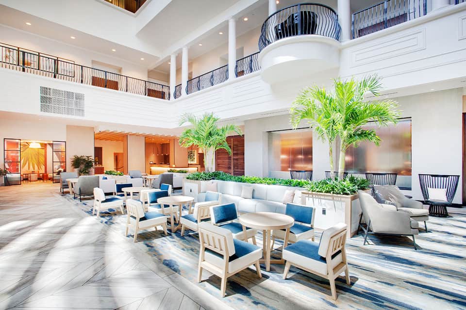 Spacious hotel lobby with high ceilings, potted palms, light wood seating, and blue accents
