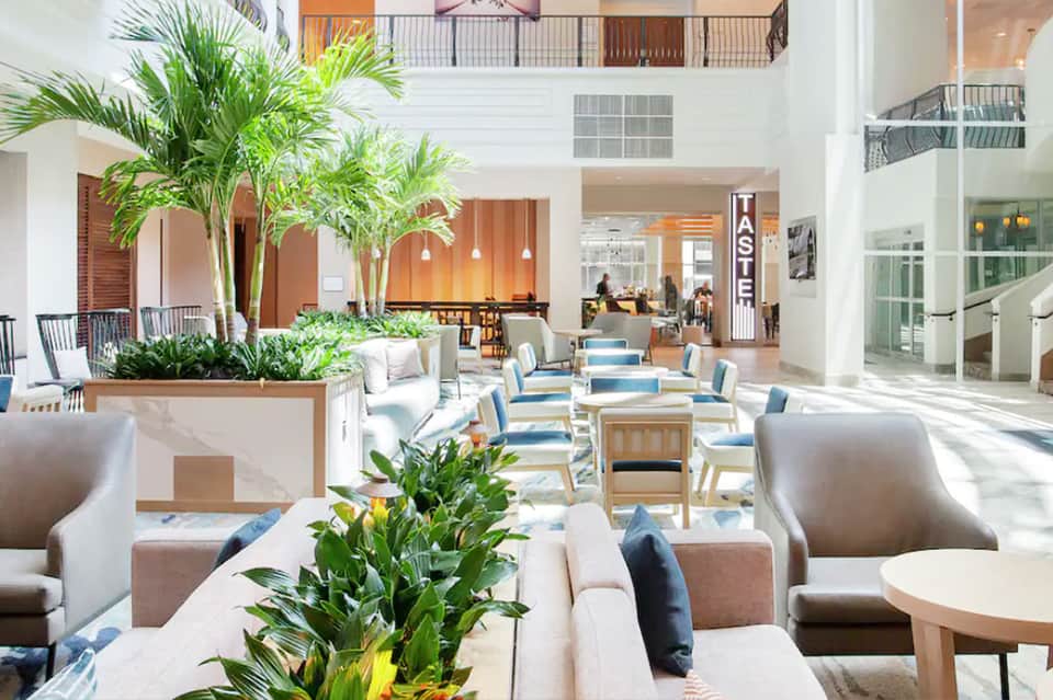 Modern hotel atrium lobby with high ceilings, palm plants, seating areas, and natural light