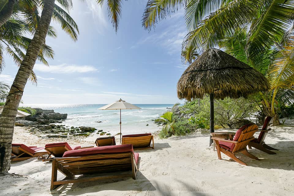 Tropical beach scene with wooden lounge chairs, thatched umbrellas, and palm trees along clear turquoise water