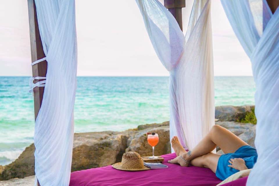 Person relaxing on pink lounger under white canopy with tropical ocean view and rocky coastline