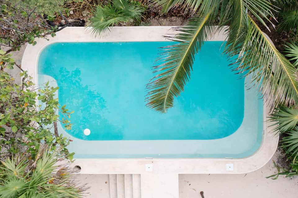 Overhead view of a turquoise swimming pool surrounded by white decking and tropical palm plants
