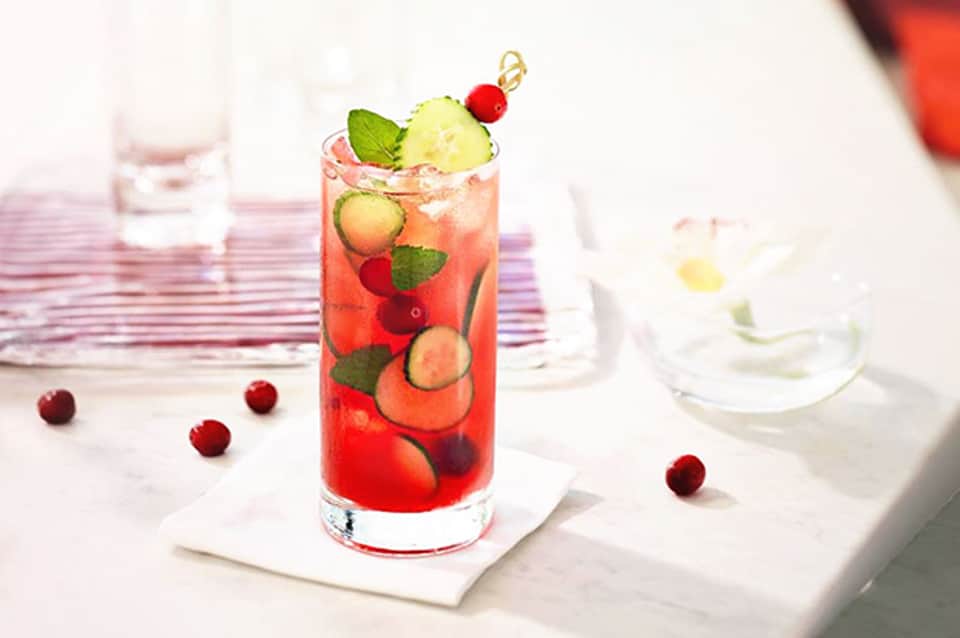 Colorful cocktail with red berries, lime, and mint garnish on white coaster at outdoor dining table