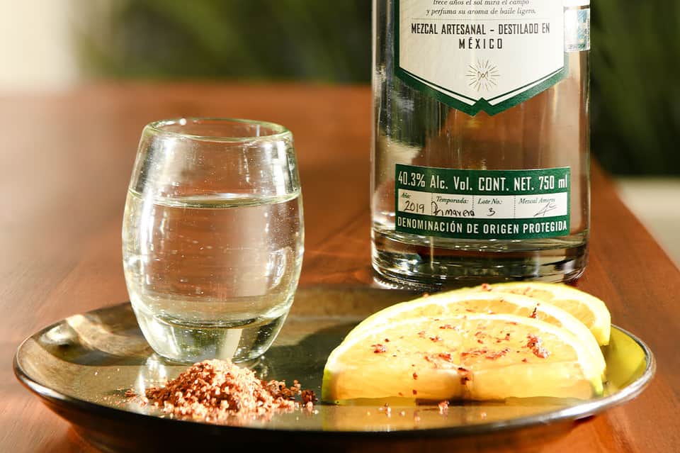 Artisanal mezcal bottle and glass with lime and salt on wooden table in Mexican sunshine