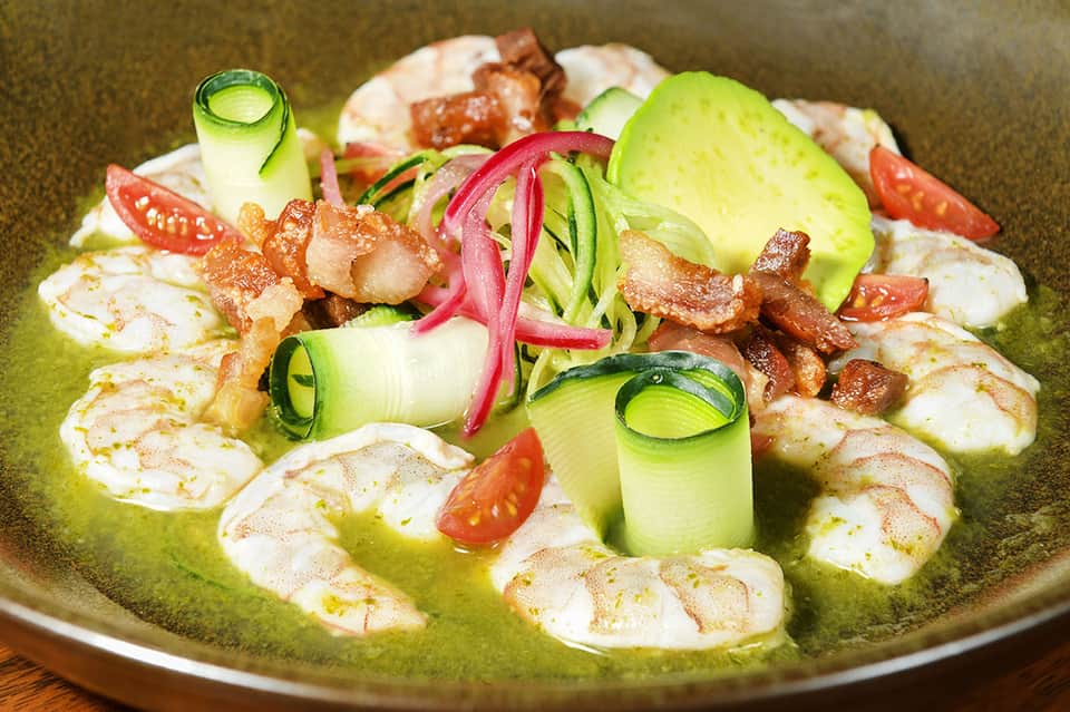 Gourmet shrimp dish plated in green sauce with cucumber and vegetable garnishes