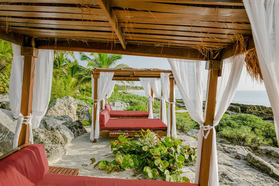 Beachfront cabana with white curtains, wooden pergola, and ocean view from sandy cove
