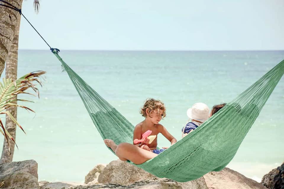 Children relaxing in green hammock strung between palm trees overlooking turquoise ocean waters