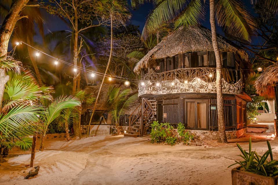 Thatched-roof bungalow illuminated at night with string lights, palm trees, and sandy beach setting