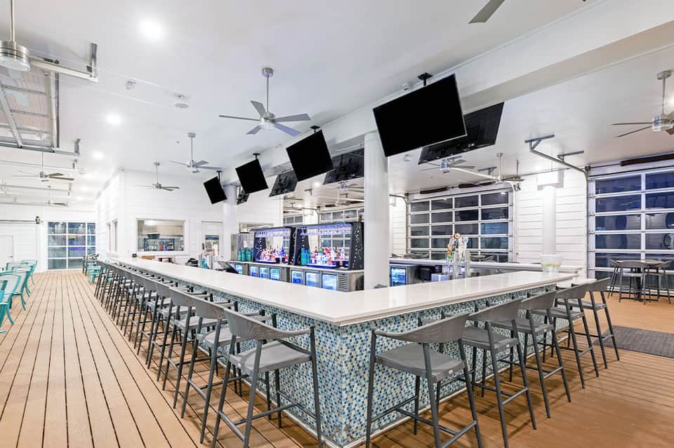 Modern resort bar with white counter, bar seating, multiple TV screens, and ocean view doors