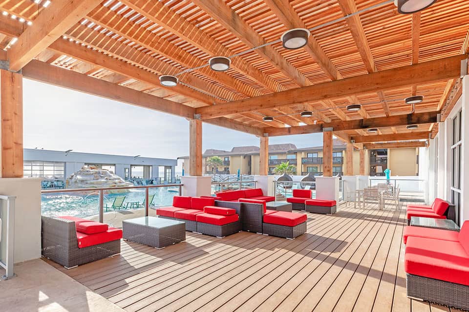 Modern poolside lounge with wooden pergola, red cushioned seating, and ocean views with palm trees