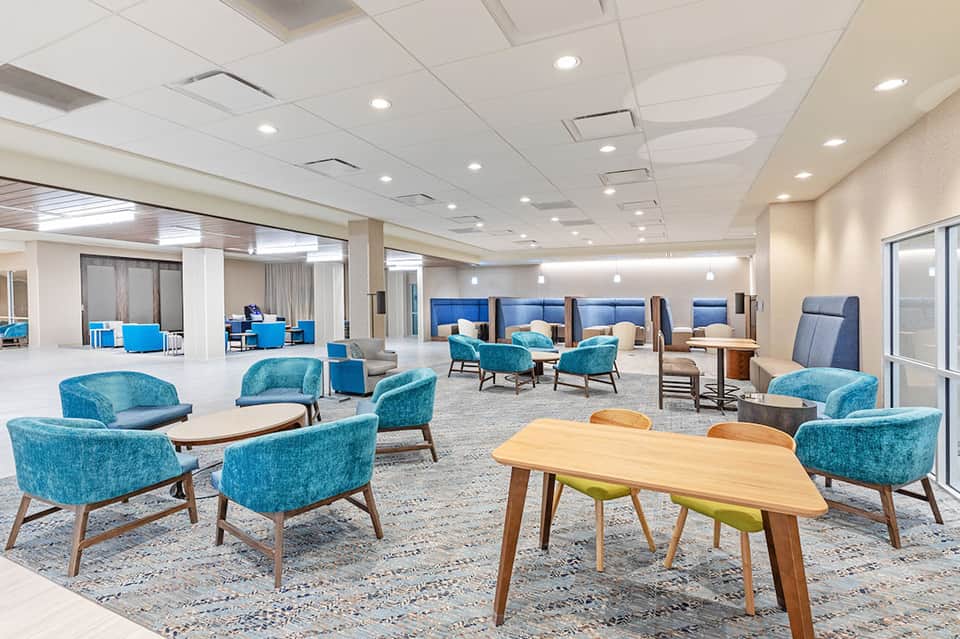 Spacious communal lounge with turquoise accent chairs, wooden tables, and modern furnishings with recessed ceiling lights