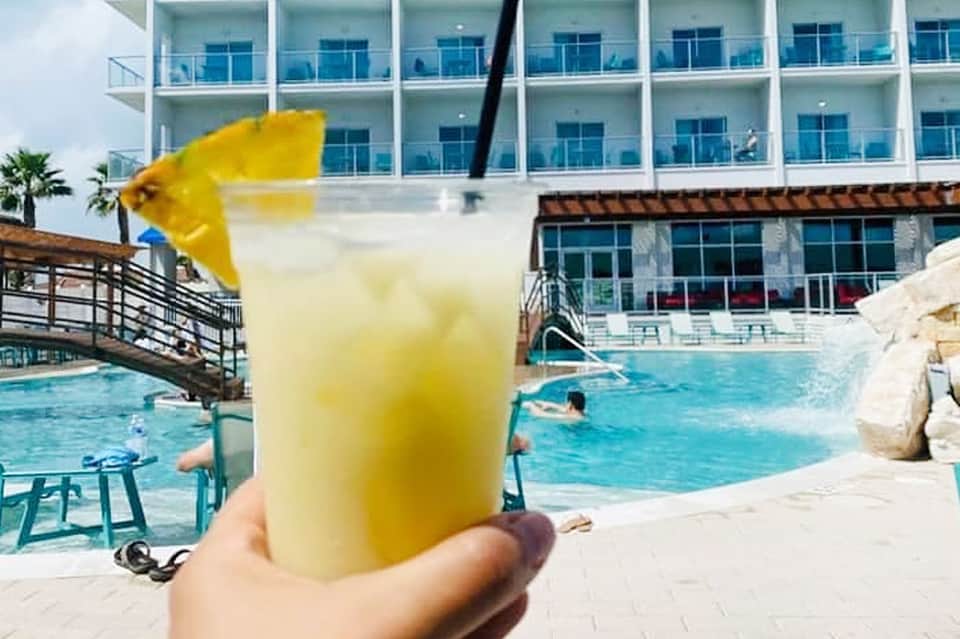 Hand holding yellow tropical drink by resort pool with modern hotel building backdrop
