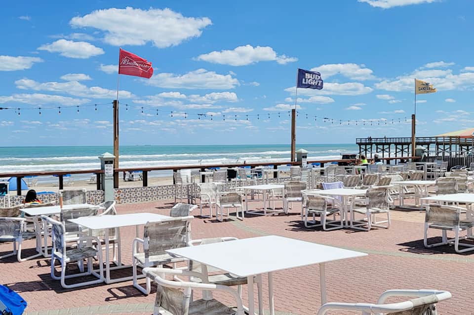 Beachfront dining area with white tables and chairs, ocean view, pier, and blue flag umbrellas