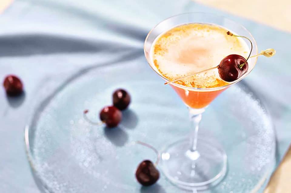 Cocktail with cherry garnish and frosted rim on blue plate with scattered berries