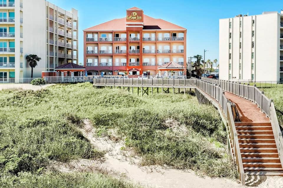 Beachfront hotel with red exterior, wooden boardwalk over dunes, and adjacent modern buildings