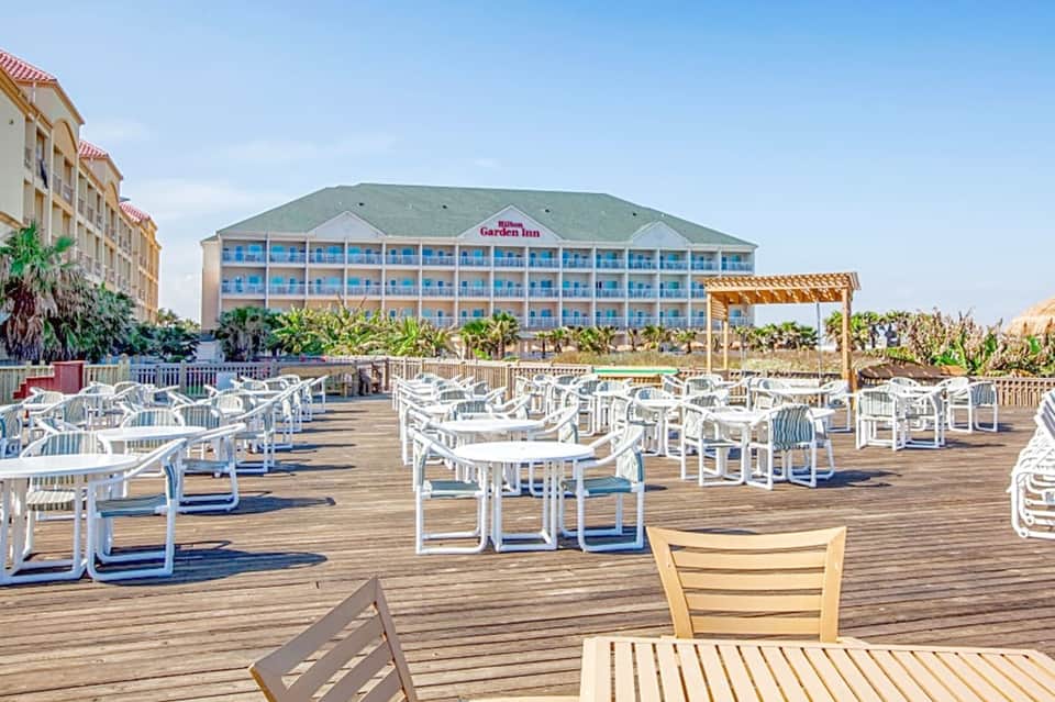 Beachfront deck with white lounge chairs and tables facing Hilton Garden Inn resort building