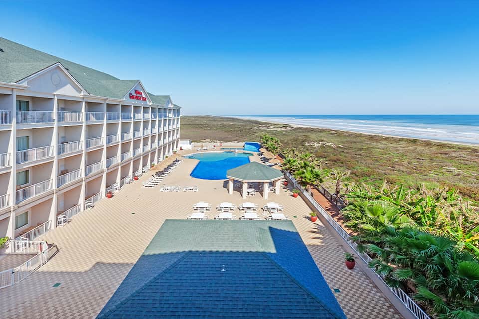 Beachfront resort with multi-story white building, blue pool, and ocean view beyond sandy dunes