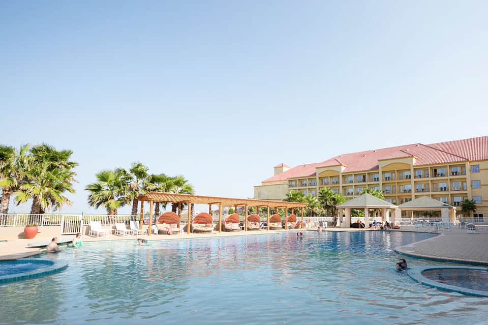 Resort pool with red umbrellas, covered cabanas, and beachfront hotel building