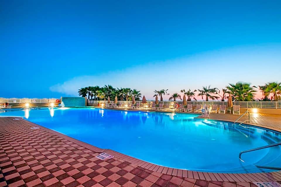 Large illuminated swimming pool at night with palm trees, lounge chairs, and resort buildings in background