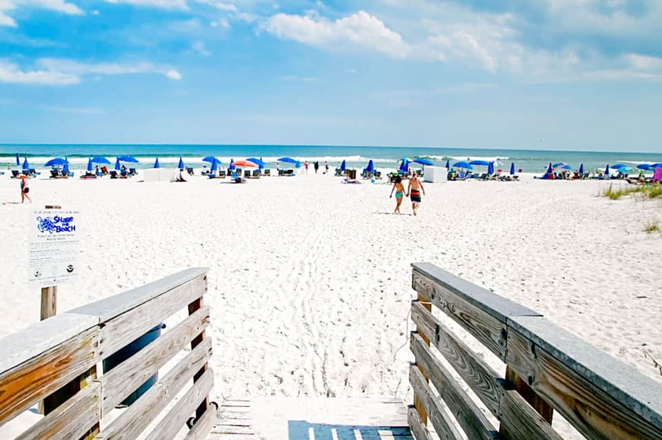 Sandy beach access with wooden boardwalk, blue umbrellas, and ocean under clear sky