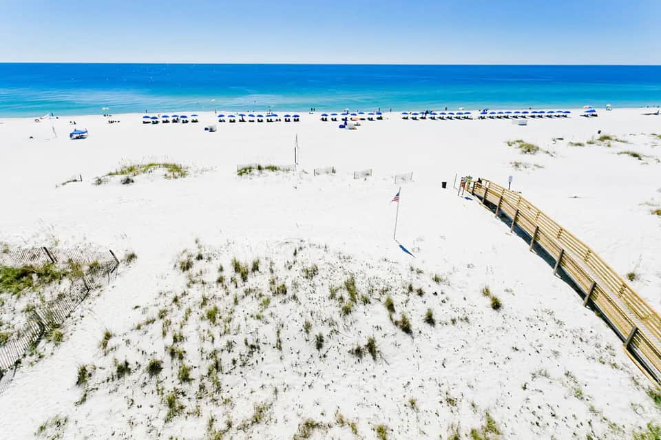 Wide sandy beach with blue umbrellas, wooden beach access fence, and clear turquoise ocean