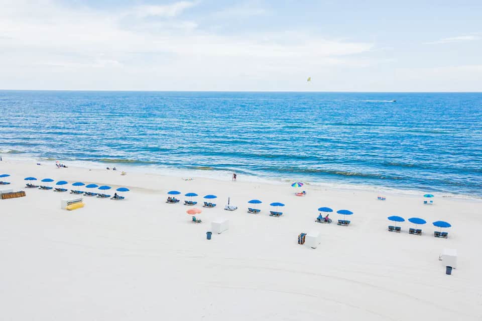 Aerial view of sandy beach with blue umbrellas, lounge chairs, and calm ocean waves