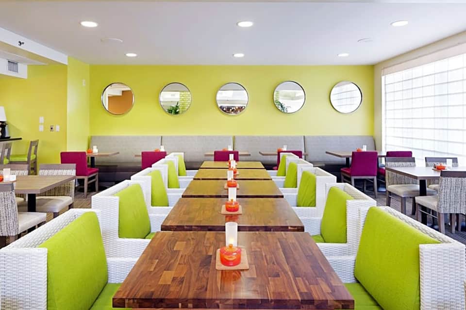 Bright yellow dining room with long wooden tables, lime green chairs, and circular mirrors on the wall