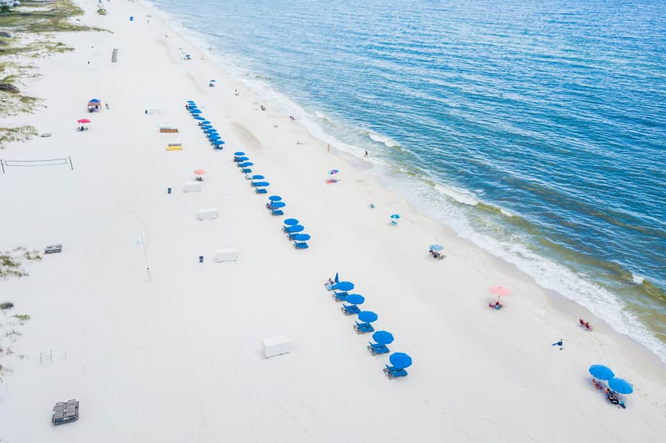 Aerial view of white sandy beach with organized rows of blue umbrellas and beach chairs along the shoreline