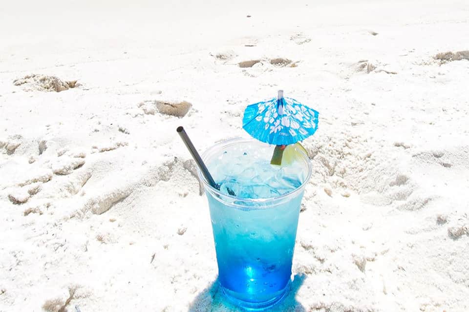 Tropical blue cocktail with umbrella and straw on sandy beach with footprints and ocean backdrop