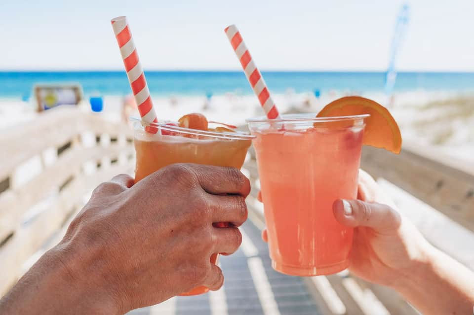 Two tropical cocktails with red and orange juices and striped straws held at beach